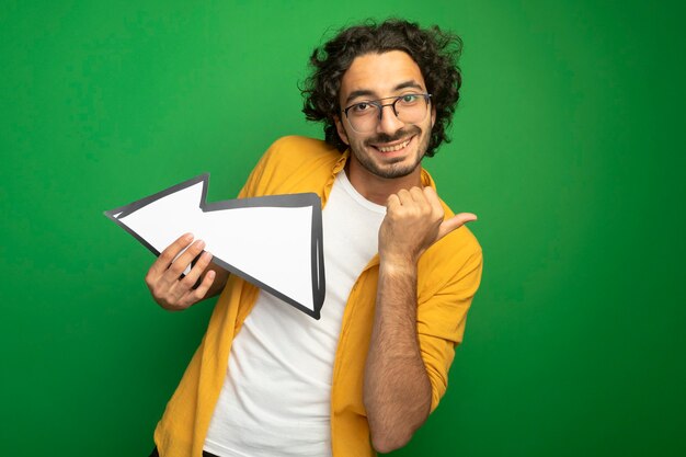 Sonriente joven apuesto hombre caucásico con gafas sosteniendo la marca de la flecha que apunta al lado mirando a la cámara apuntando al lado aislado sobre fondo verde con espacio de copia