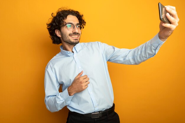 Sonriente joven apuesto hombre caucásico con gafas tomando selfie aislado sobre fondo naranja con espacio de copia