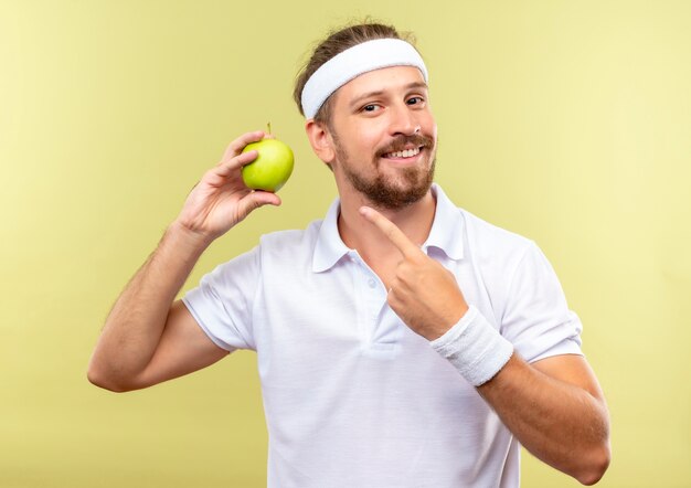 Sonriente joven apuesto hombre deportivo con diadema y muñequeras sosteniendo y apuntando a apple aislado en espacio verde