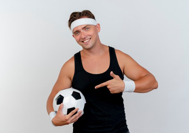 Sonriente joven apuesto hombre deportivo con diadema y muñequeras sosteniendo y apuntando a un balón de fútbol aislado en la pared blanca