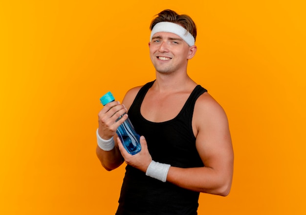 Sonriente joven apuesto hombre deportivo con diadema y muñequeras sosteniendo una botella de agua aislada en la pared naranja