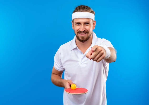 Sonriente joven apuesto hombre deportivo con diadema y muñequeras sosteniendo la raqueta de ping pong con pelota y apuntando aislado en el espacio azul