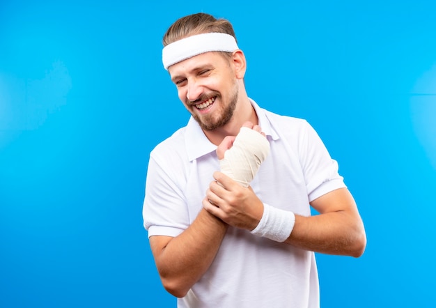 Sonriente joven apuesto hombre deportivo con diadema y muñequeras sosteniendo su muñeca lesionada envuelta con una venda aislada en el espacio azul