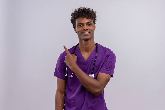 Un sonriente joven apuesto médico de piel oscura con cabello rizado vistiendo uniforme violeta con estetoscopio apuntando hacia arriba mientras