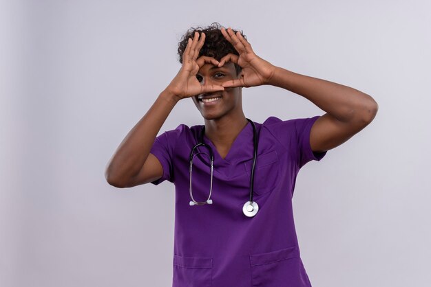 Un sonriente joven apuesto médico de piel oscura con cabello rizado vistiendo uniforme violeta con estetoscopio mostrando signo de forma de corazón con los dedos