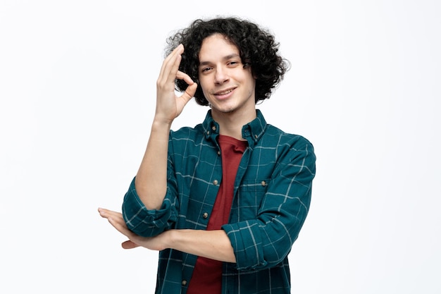 Foto gratuita sonriente joven apuesto mirando a la cámara manteniendo la mano bajo el codo haciendo un buen signo aislado en el fondo blanco