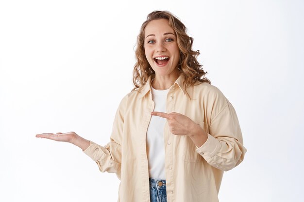 Sonriente joven apuntando a su mano con su producto, exhiba el artículo en su palma contra el espacio de la copia, de pie sobre una pared blanca