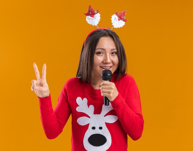 Sonriente joven asiática con aro de pelo de navidad con suéter habla en el micrófono mostrando gesto de paz aislado en la pared naranja