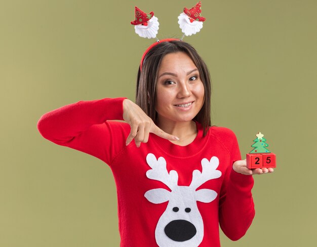 Sonriente joven asiática vistiendo el aro de pelo de Navidad sosteniendo y puntos en el juguete de Navidad aislado sobre fondo verde oliva