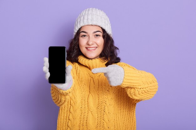 Sonriente joven atractiva mujer apuntando a la pantalla en blanco del teléfono en su mano