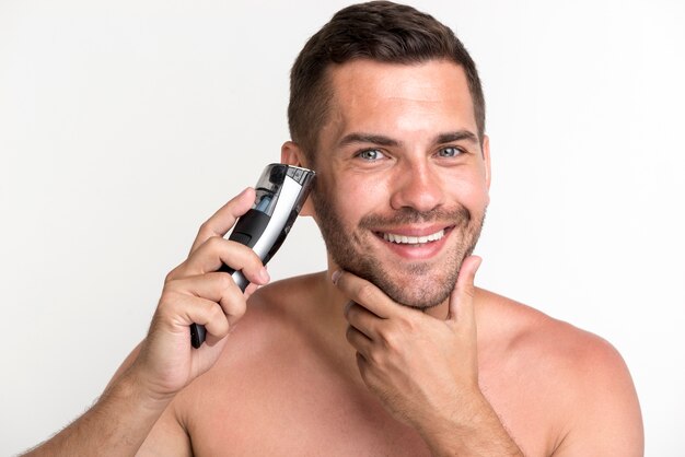 Sonriente joven barba de afeitar con máquina de afeitar eléctrica sobre fondo blanco.