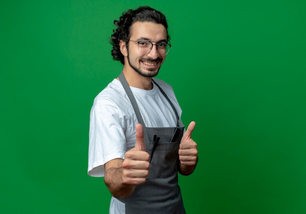 Sonriente joven barbero con gafas y banda de pelo ondulado en uniforme mostrando los pulgares para arriba