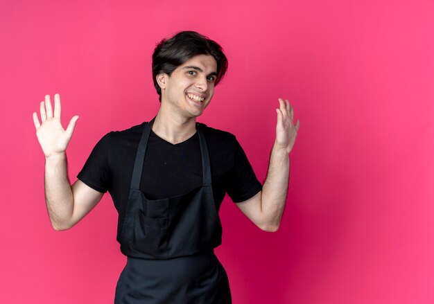 Sonriente joven barbero guapo en uniforme extiende las manos aisladas en rosa con espacio de copia