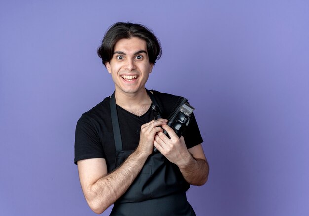 Sonriente joven barbero guapo en uniforme sosteniendo barbero thools aislado en azul