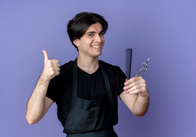 Sonriente joven barbero guapo en uniforme sosteniendo el peine con tijeras y su pulgar hacia arriba aislado en azul