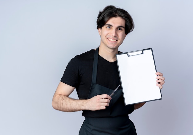 Sonriente joven barbero guapo en uniforme sosteniendo portapapeles con tijeras aislado en blanco