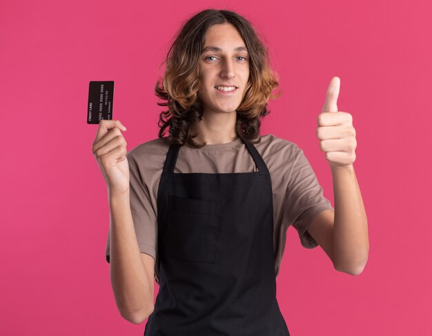 Sonriente joven barbero guapo vistiendo uniforme mostrando tarjeta de crédito y pulgar hacia arriba