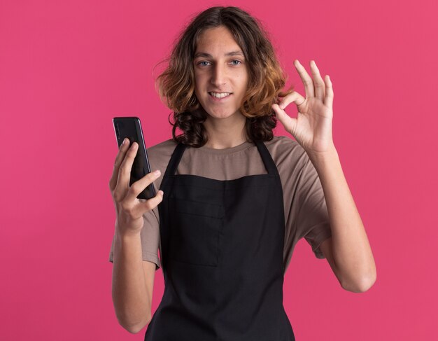 Sonriente joven barbero guapo vistiendo uniforme sosteniendo teléfono móvil haciendo bien signo aislado en pared rosa