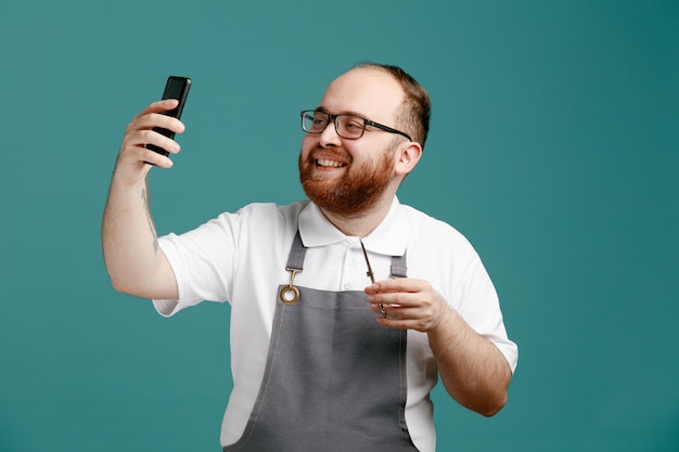 Sonriente joven barbero con uniforme y gafas sosteniendo tijeras tomando selfie con teléfono móvil aislado sobre fondo azul.