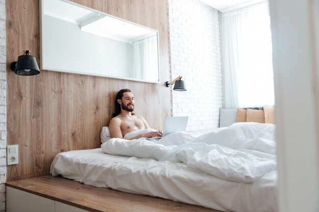 Sonriente joven barbudo sentado y usando la computadora portátil en la cama