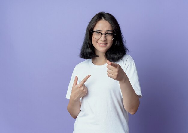 Sonriente joven bastante caucásica con gafas apuntando a sí misma y a la cámara aislada sobre fondo púrpura con espacio de copia