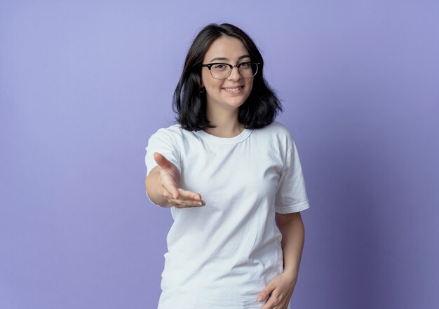 Sonriente joven bastante caucásica con gafas estirando la mano a la cámara aislada sobre fondo púrpura con espacio de copia