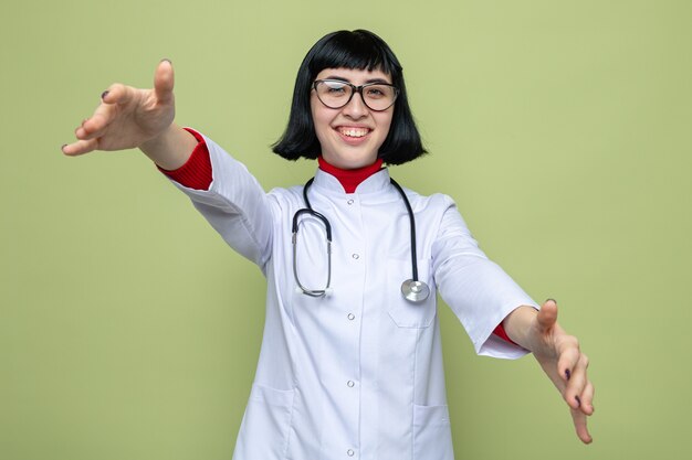 Sonriente joven bastante caucásica con gafas ópticas en uniforme médico con estetoscopio estirando las manos