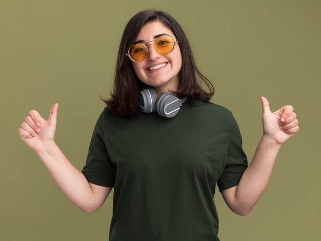 Sonriente joven bastante caucásica en gafas de sol y con auriculares alrededor del cuello pulgares arriba aislado en la pared verde oliva con espacio de copia