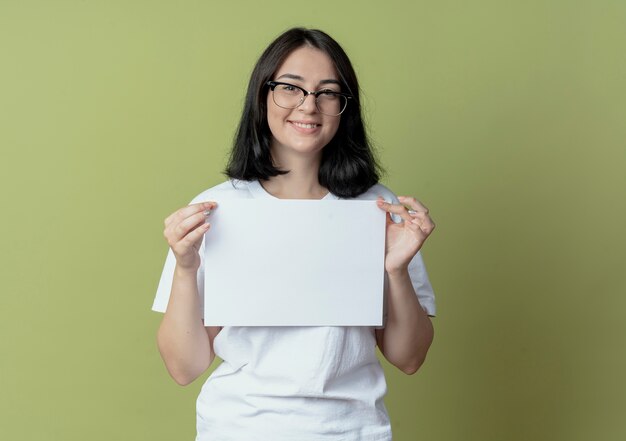 Sonriente joven bastante caucásica con gafas sosteniendo papel aislado sobre fondo verde oliva con espacio de copia