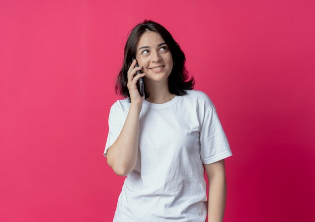 Sonriente joven bastante caucásica hablando por teléfono y mirando al lado aislado sobre fondo carmesí con espacio de copia