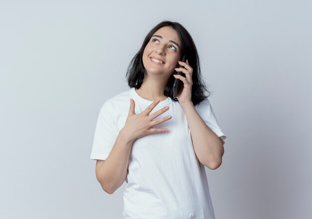 Sonriente joven bastante caucásica hablando por teléfono y mirando hacia arriba aislado sobre fondo blanco con espacio de copia