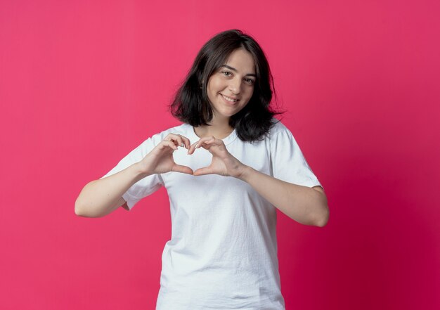 Sonriente joven bastante caucásica haciendo signo de corazón en cámara aislada sobre fondo carmesí con espacio de copia