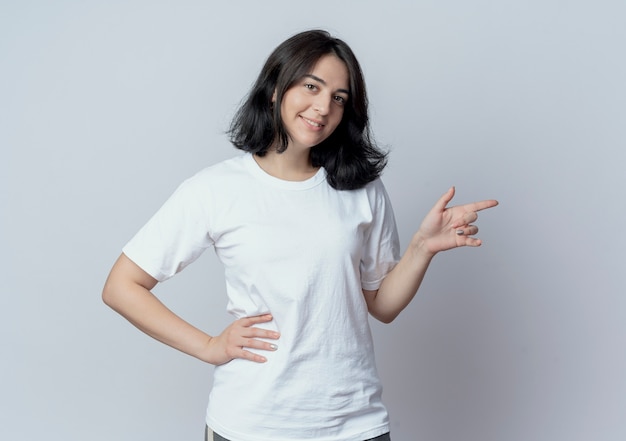 Sonriente joven bastante caucásica manteniendo la mano en la cintura y apuntando al lado aislado sobre fondo blanco con espacio de copia