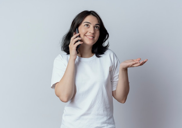 Sonriente joven bastante caucásica mirando hacia arriba hablando por teléfono y mostrando la mano vacía aislada sobre fondo blanco con espacio de copia