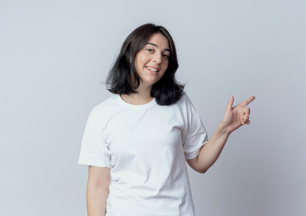 Sonriente joven bastante caucásica mirando a la cámara y apuntando al lado aislado sobre fondo blanco con espacio de copia