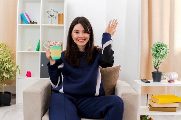 Sonriente joven bastante caucásica sentada en un sillón en la sala de estar diseñada sosteniendo la taza manteniendo la mano en el aire