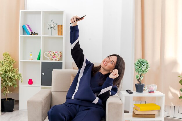 Sonriente joven bastante caucásica sentada en un sillón en la sala de estar diseñada tomando selfie y manteniendo la mano en el aire