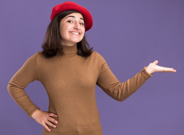 Sonriente joven bastante caucásica con sombrero boina apuntando al lado con la mano