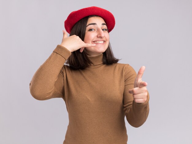 Sonriente joven bastante caucásica con sombrero de boina gesticulando llámame firmar y apuntando a la cámara