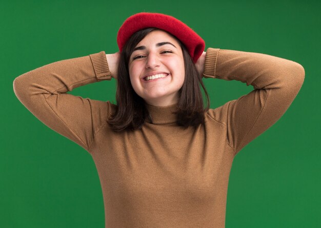 Sonriente joven bastante caucásica con sombrero de boina pone las manos en la cabeza detrás aislado en la pared verde con espacio de copia