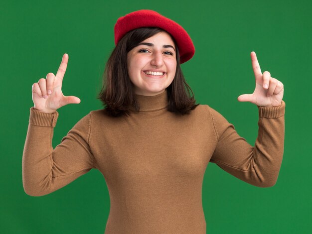 Sonriente joven bastante caucásica con sombrero de boina que gesticula el marco con las manos aisladas en la pared verde con espacio de copia