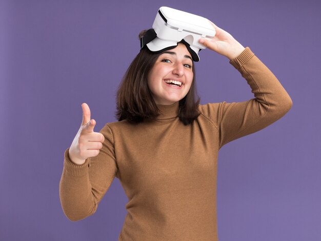 Sonriente joven bastante caucásica con sombrero de boina sosteniendo auriculares vr y apuntando a la cámara en púrpura