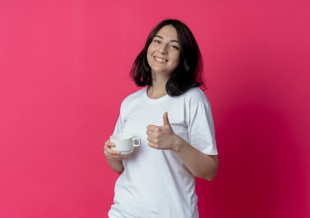 Sonriente joven bastante caucásica sosteniendo la taza y mostrando el pulgar hacia arriba aislado sobre fondo carmesí con espacio de copia