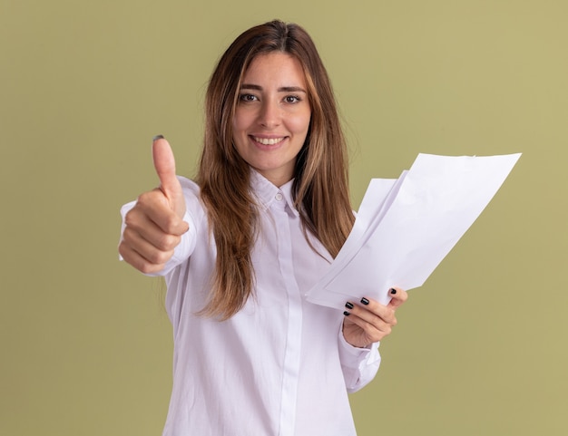 Sonriente joven bastante caucásica sostiene hojas de papel en blanco y pulgares arriba aislados en la pared verde oliva con espacio de copia