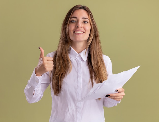 Foto gratuita sonriente joven bastante caucásica thumbs up y sostiene hojas de papel en blanco