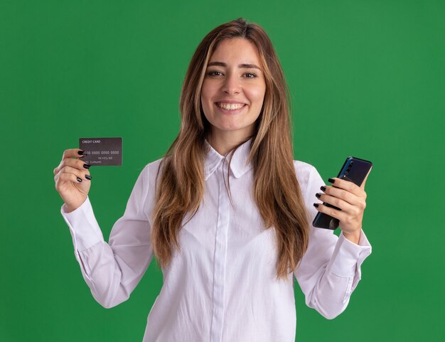 Sonriente joven bastante caucásica tiene tarjeta de crédito y teléfono en verde