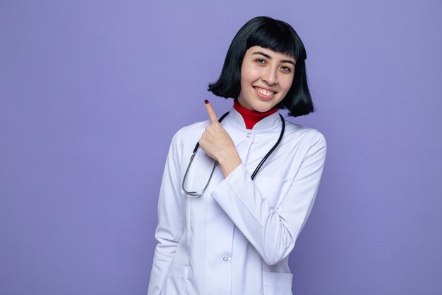 Sonriente joven bastante caucásica en uniforme médico con estetoscopio apuntando hacia arriba