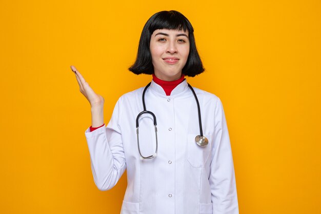 Sonriente joven bastante caucásica en uniforme médico con estetoscopio manteniendo la mano abierta