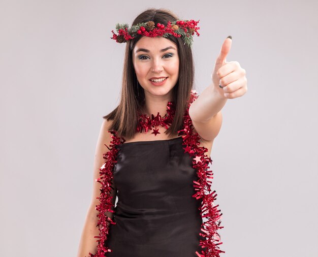 Sonriente joven bastante caucásica vestida con corona de Navidad y guirnalda de oropel alrededor del cuello mirando a cámara mostrando el pulgar hacia arriba aislado sobre fondo blanco con espacio de copia