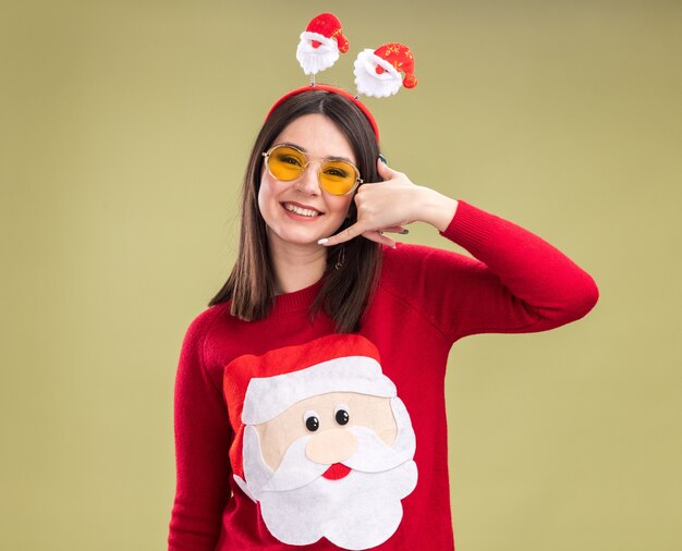 Sonriente joven bastante caucásica vistiendo suéter de santa claus y diadema con gafas mirando a cámara haciendo gesto de llamada aislado sobre fondo verde oliva